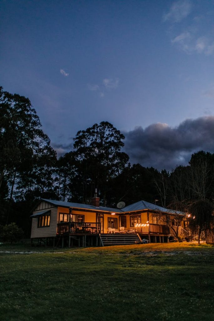 Facade of illuminated residential building located on grassy terrain near tall trees against dark cloudy sky in countryside during dusk time